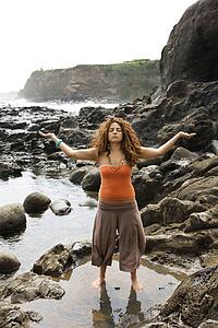 Young adult female standing on rocky shore with arms outstretched and eyes closed.