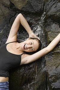 Caucasian young adult woman standing under small fresh waterfall laughing with arms over head and looking at viewer.