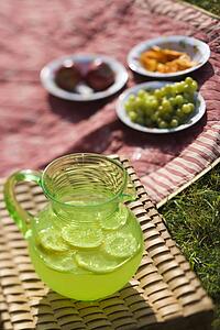 Green plastic pitcher of lemonade and lemons with a picnic spread out in the background