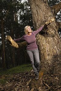 Middle-aged Caucasian woman smiling standing holding tree in forest.
