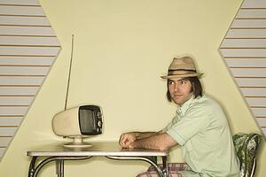 Caucasian mid-adult man wearing hat sitting at 50's retro dinette set in front of old television.