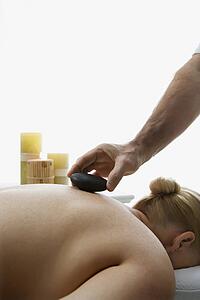 Male massage therapist placing hot stone on back of woman laying on massage table