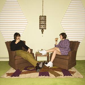 Caucasian mid-adult man and woman wearing vintage clothing seated in brown retro chairs smiling and drinking.