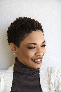Close up head and shoulder of African-American woman standing against white wall smiling with head turned to side.