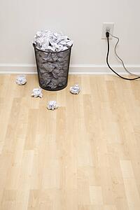 Full wire mesh trash can with crumpled paper next to electrical outlet and plugs