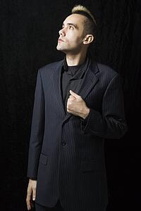 Caucasian man in suit with mohawk standing posed against black background.