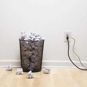 Full wire mesh trash can with crumpled paper next to electrical outlet and plugs