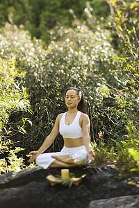 Asian American woman sitting on boulder in forest meditating with candle in Maui, Hawaii.