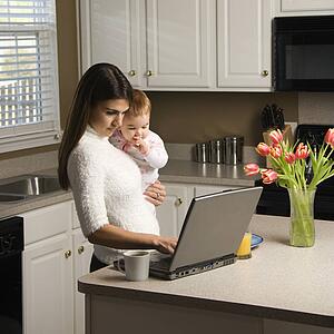 Woman holding baby and typing on laptop computer in kitchen