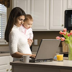 Mother holding baby and typing on laptop computer in kitchen