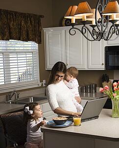 Mother holding baby and typing on laptop computer with girl eating breakfast in kitchen