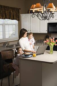 Mother holding baby and typing on laptop computer with girl eating breakfast in kitchen