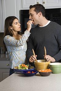 Woman feeding man at kitchen counter while he makes salad