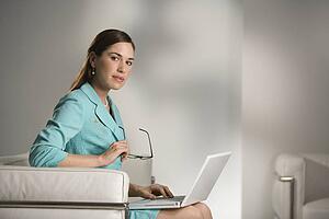Professional business woman sitting in modern office working on laptop computer holding eyeglasses and looking at viewer
