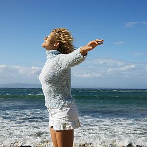 Pretty young blond woman standing on Maui, Hawaii beach with arms out to side and wind blowing hair smiling.
