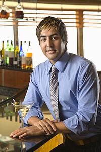 Portrait of Hispanic mid adult man leaning on bar with martini smiling at viewer.