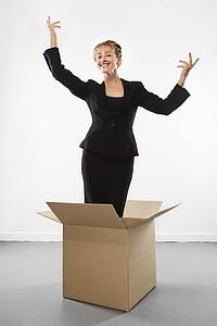 Businesswoman standing in cardboard box gesturing above head and smiling at viewer