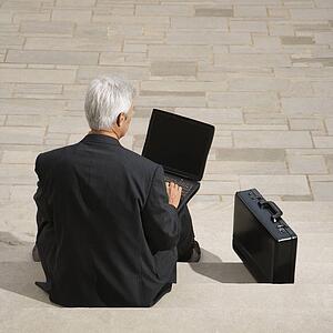 Back view of middle aged businessman sitting on steps outdoors with laptop and briefcase