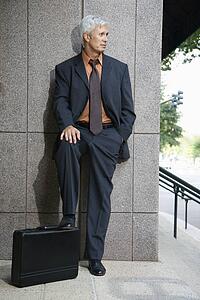 Caucasian middle aged businessman leaning on wall outdoors with foot on briefcase.