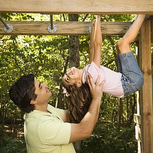 Hispanic girl hanging by arms and legs from monkey bars smiling at father helping her.