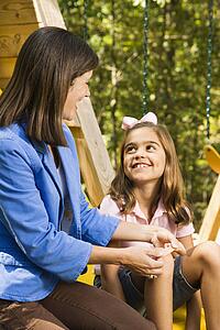 Hispanic girl sitting on playground slide smiling at woman applying first aid bandage to knee.