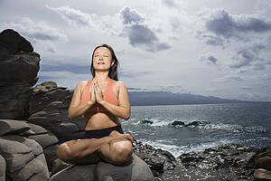 Asian woman sitting on rock by ocean in lotus pose with eyes closed in Maui, Hawaii