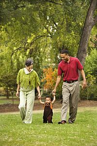 Mother and father helping toddler walk holding his hands in park.