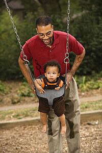 Father pushing young son on swing in park in playground
