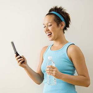 Young woman in fitness clothing holding bottled water and smiling at cellphone.
