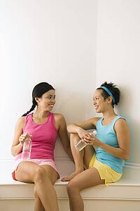 Two young women in fitness clothes holding water bottles sitting smiling and talking.