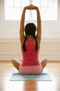 Rear view of young woman sitting on mat with legs crossed stretching.