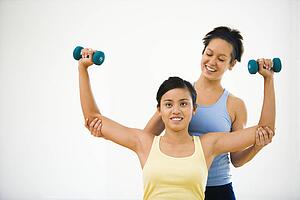 Young woman lifting hand weights while another woman helps position her arms