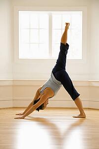 Young woman doing downward dog pose with one leg raised on wooden floor indoors by sunlit window.