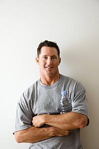 Head and shoulder portrait of man in t-shirt against white wall holding water bottle and smiling.