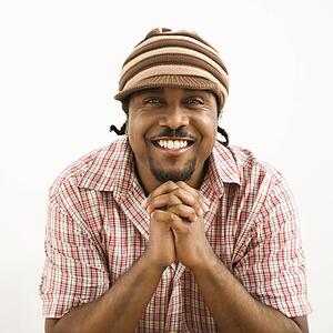 African-American mid-adult man wearing hat and smiling at viewer with hands together.