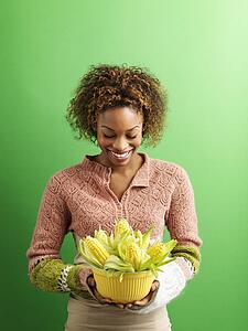 Portrait of pretty young woman standing against green background holding bowl full of ears of corn.