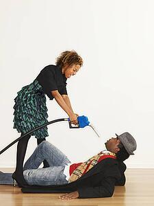 Man on floor with angry woman standing over him pointing gasoline pump nozzle at him like a gun.