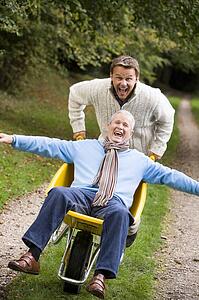 Man walking on path outdoors pushing other man in wheelbarrow and smiling (selective focus)
