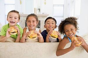 Four young children eating cheeseburgers in living room smiling