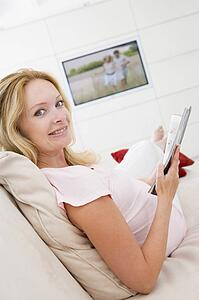 Pregnant woman reading magazine with television in background smiling