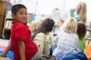 Student in class looking at camera with teacher reading in background (selective focus)