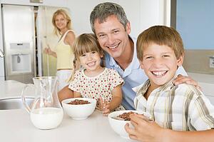 Father With Children As They Eat Breakfast And Mother In The Back Ground 