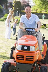 Man outdoors driving lawnmower smiling with family in background