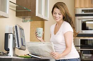 Woman in kitchen at computer with newspaper and coffee smiling