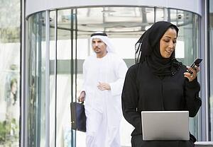 Businesswoman sitting outdoors by building with laptop holding cellular phone with businessman running in background (selective focus)
