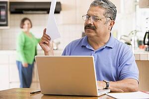 Man in kitchen with laptop and paperwork with woman in background