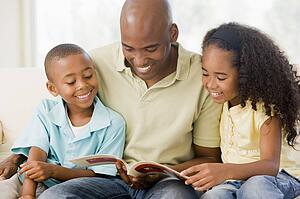 Man and two children sitting in living room reading book and smiling