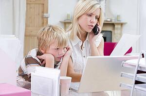 Woman using telephone in home office with laptop while young boy waits