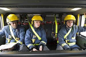 Three firefighters in fire engine wearing helmets with one reading