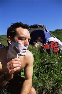 Man outdoors shaving at campsite with woman in background (selective focus)